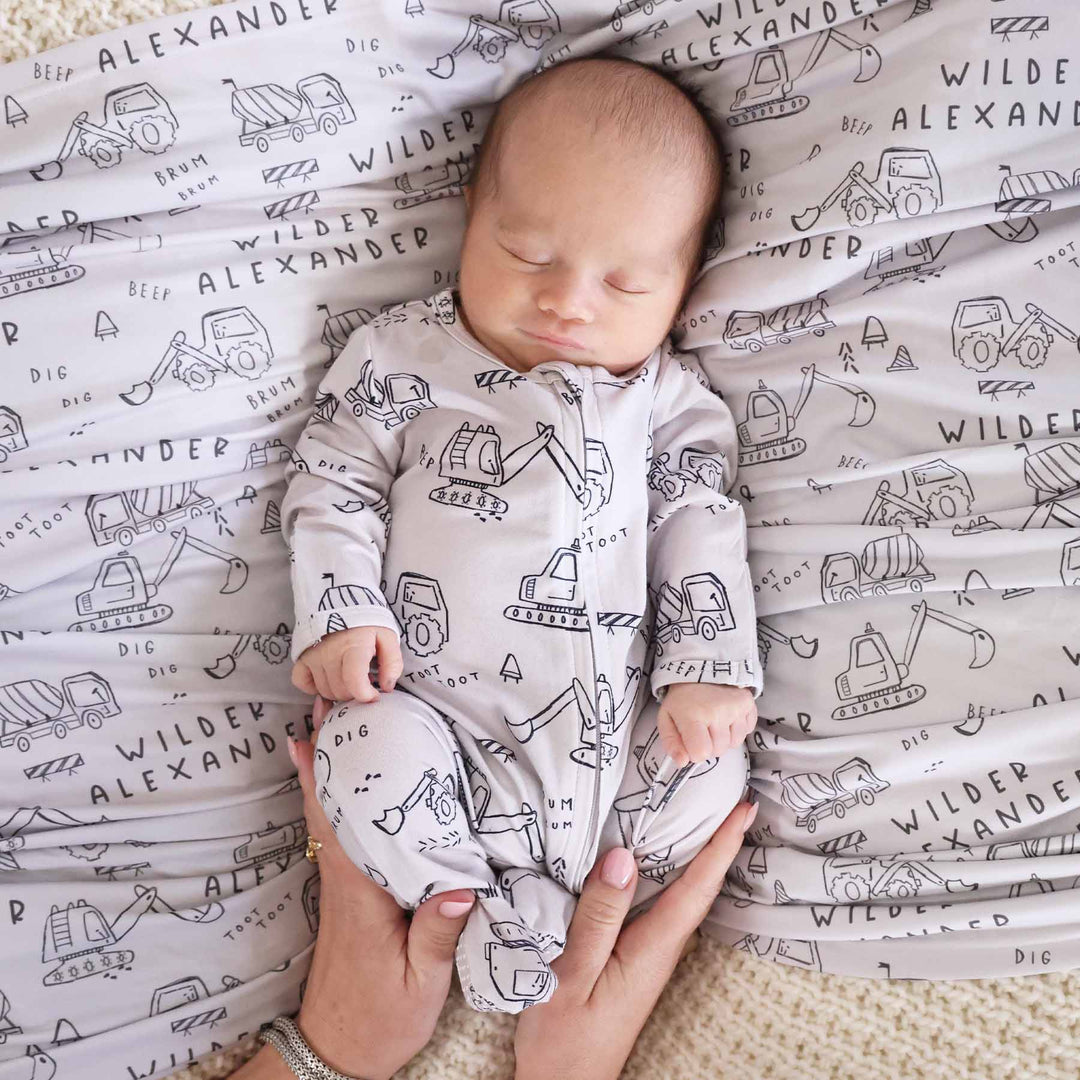 newborn baby boy with personalized swaddle in construction truck and dozer doodle print with matching zip footie romper