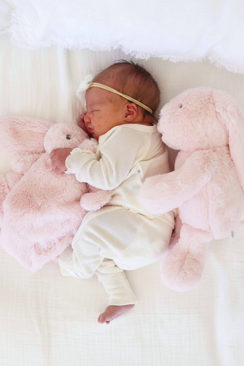 baby laying on side with pink bunny stuffed animal behind her holding a pink embroidered bunny lovey