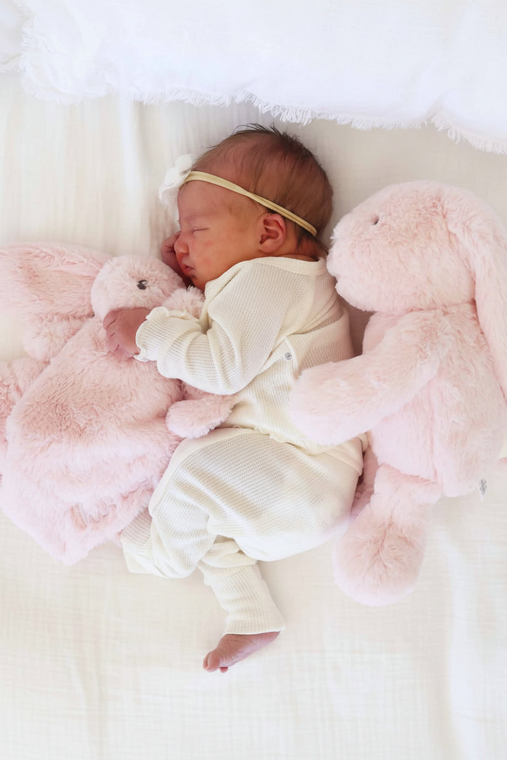 baby laying on side with pink bunny stuffed animal behind her holding a pink embroidered bunny lovey