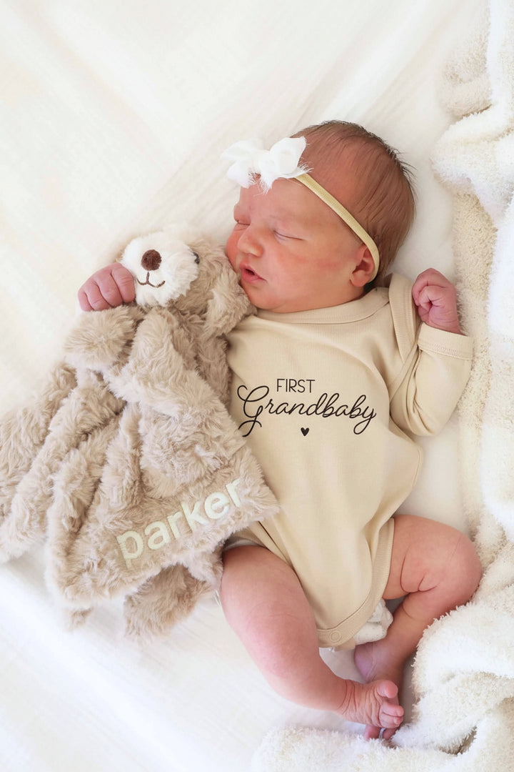 baby laying on bed in first grandbaby graphic bodysuit holding an embroidered puppy lovey