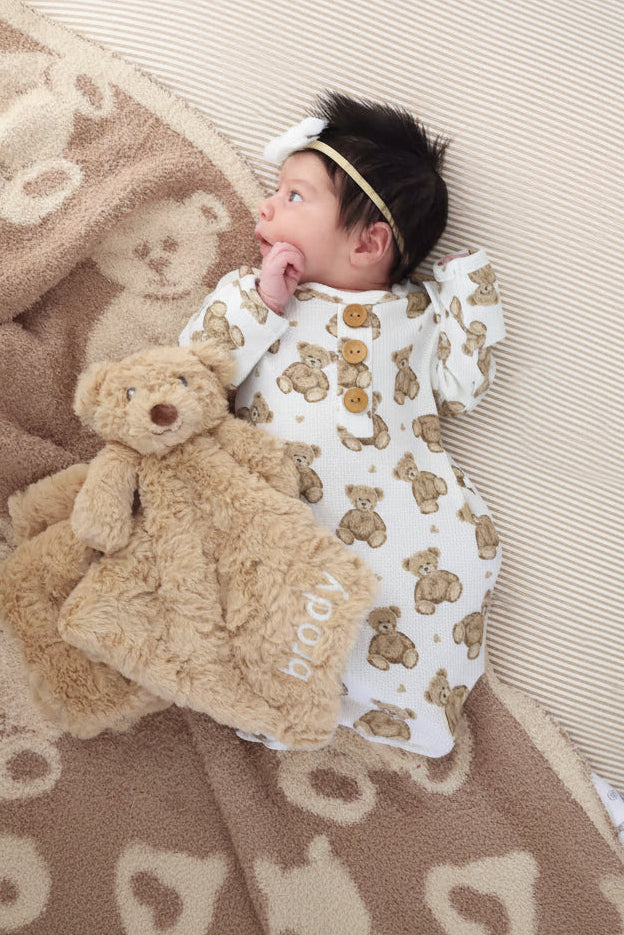 baby laying in crib with brown teddy bear embroidered lovey in bear knot gown with bear blanket and brown and white crib sheet