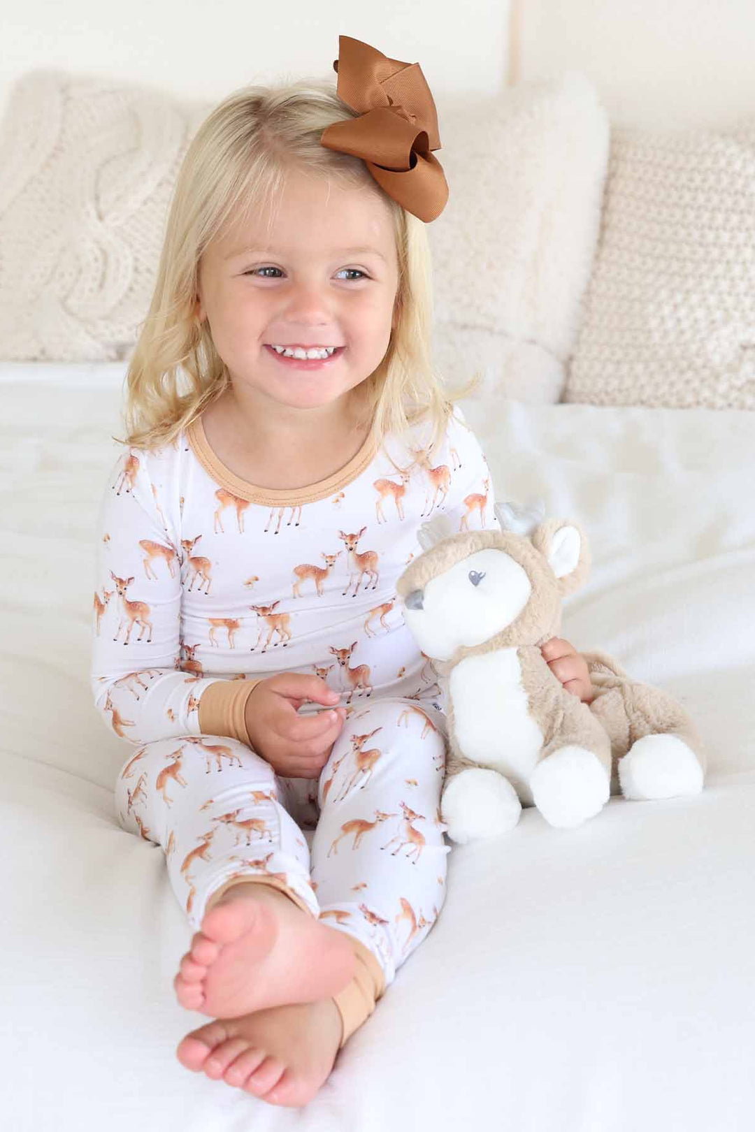 Smiling toddler in fawn print pajamas holding a plush deer on a cozy bed