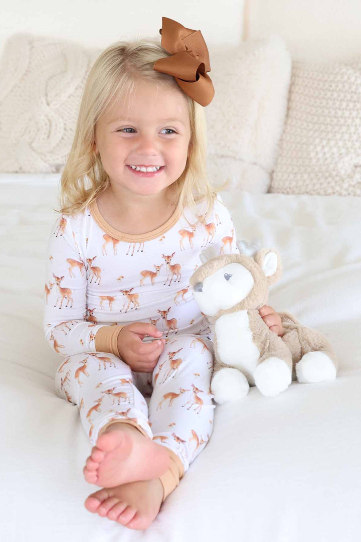 Smiling toddler in fawn print pajamas holding a plush deer on a cozy bed