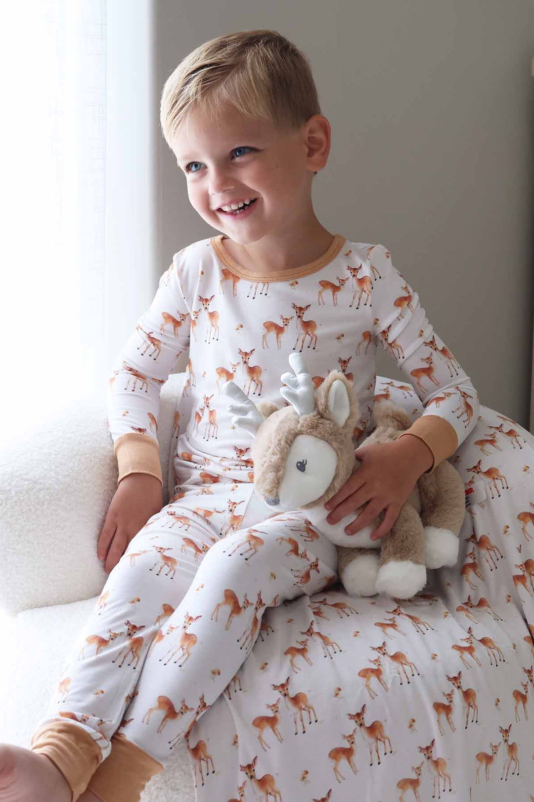 Little boy smiling in woodland fawn print pajamas beside a stuffed deer