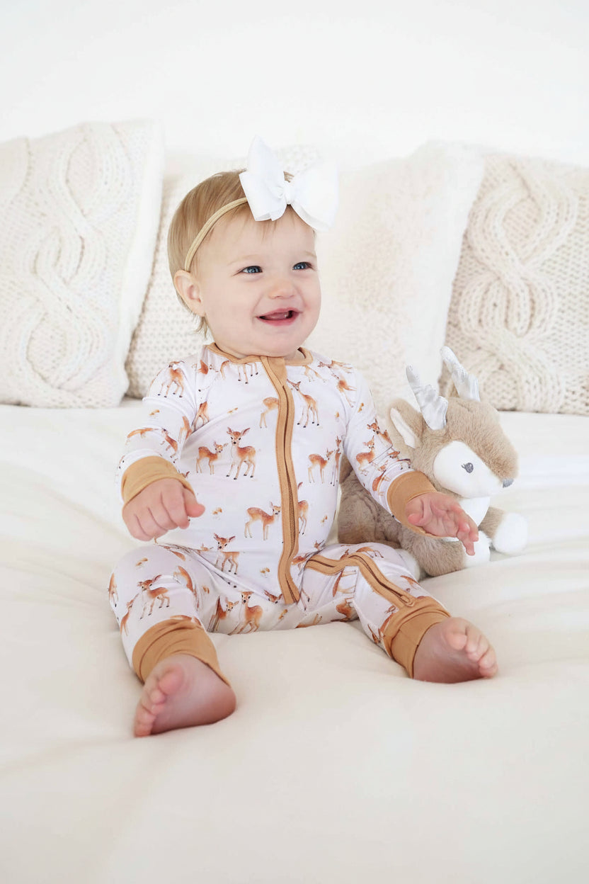baby girl sitting on bed wearing a fawn convertible zip romper pajama with a white ribbon bow headband next to a deer stuffed animal