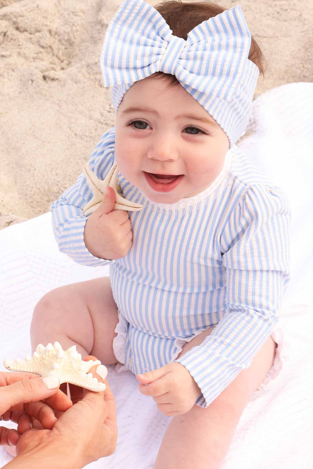 baby girl sitting on beach towel in blue seersucker long sleeve rash guard with ruffle bottom with matching swim bow