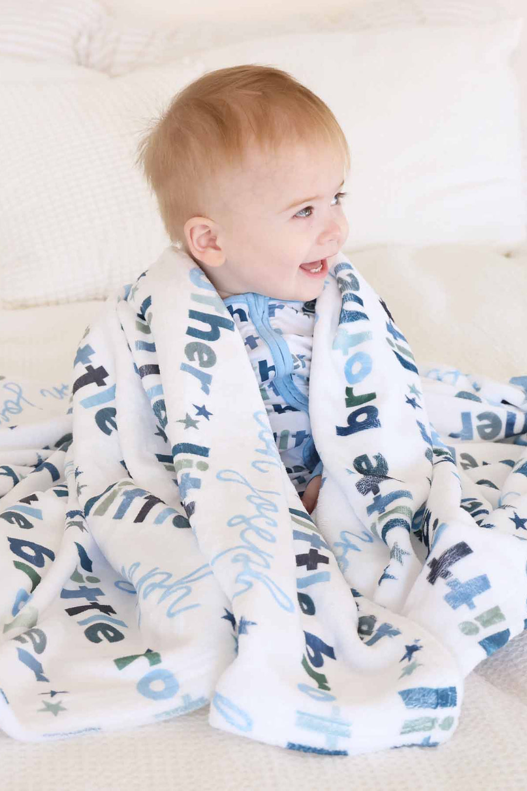 Baby sitting on bed and covering with the personalized little brother blanket. 