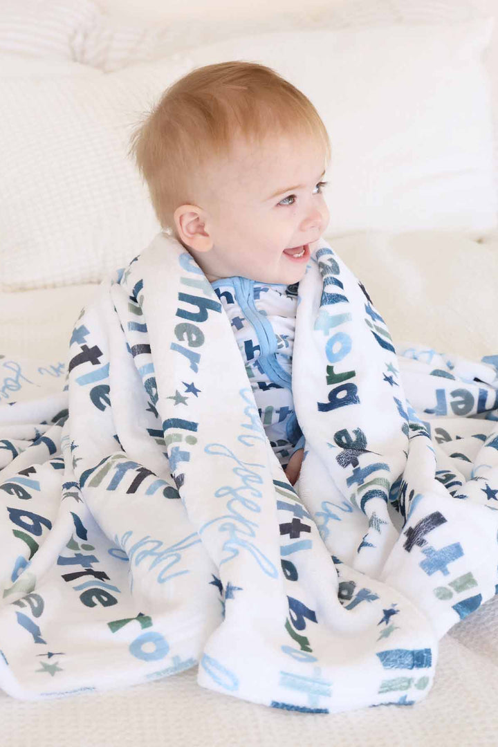 Baby sitting on bed and covering with the personalized little brother blanket. 