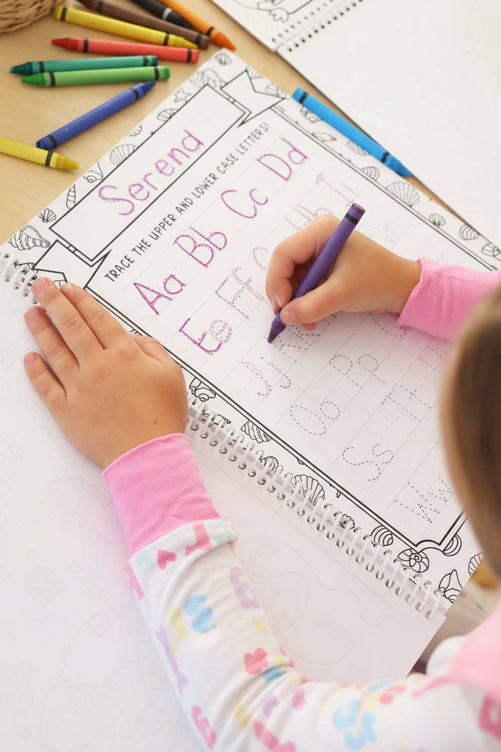 Child in a Big Sister pajama set tracing personalized 'Serena' alphabet letters with a purple crayon on a seashell-bordered practice page in a Caden Lane coloring book.
