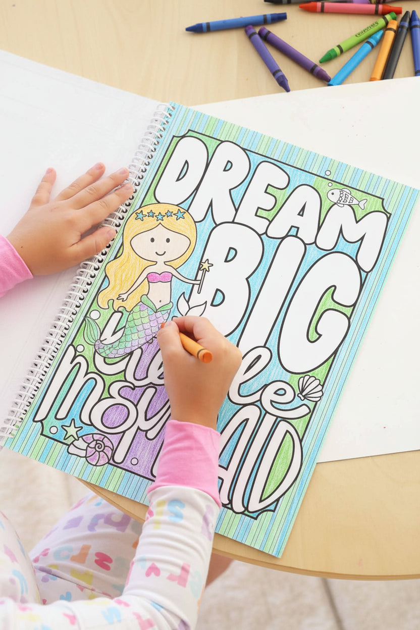 Child in a Big Sister pajama set coloring a 'Dream Big Little Mermaid' page with an orange crayon, filling in a mermaid illustration in a Caden Lane coloring book.