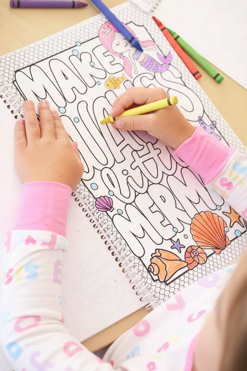 Child in a Big Sister pajama set coloring a 'Make Waves Little Mermaid' page with a yellow crayon in a Caden Lane coloring book, surrounded by seashell illustrations