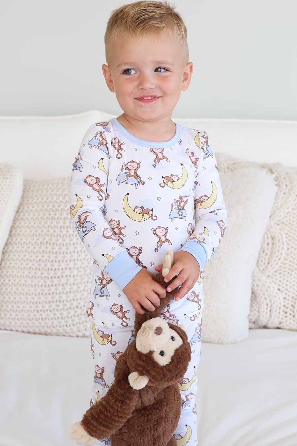 boy standing on bed wearing a two piece pajama set for kids with monkeys sleeping on banana moons and jumping on beds holding a monkey stuffed animal
