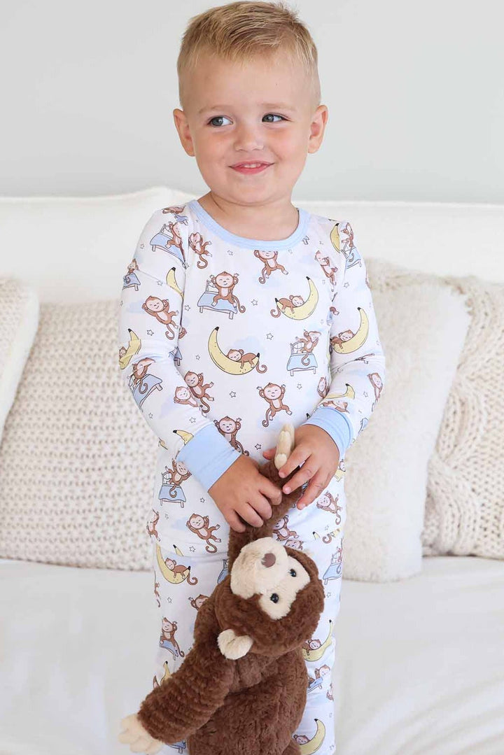 boy standing on bed wearing a two piece pajama set for kids with monkeys sleeping on banana moons and jumping on beds holding a monkey stuffed animal