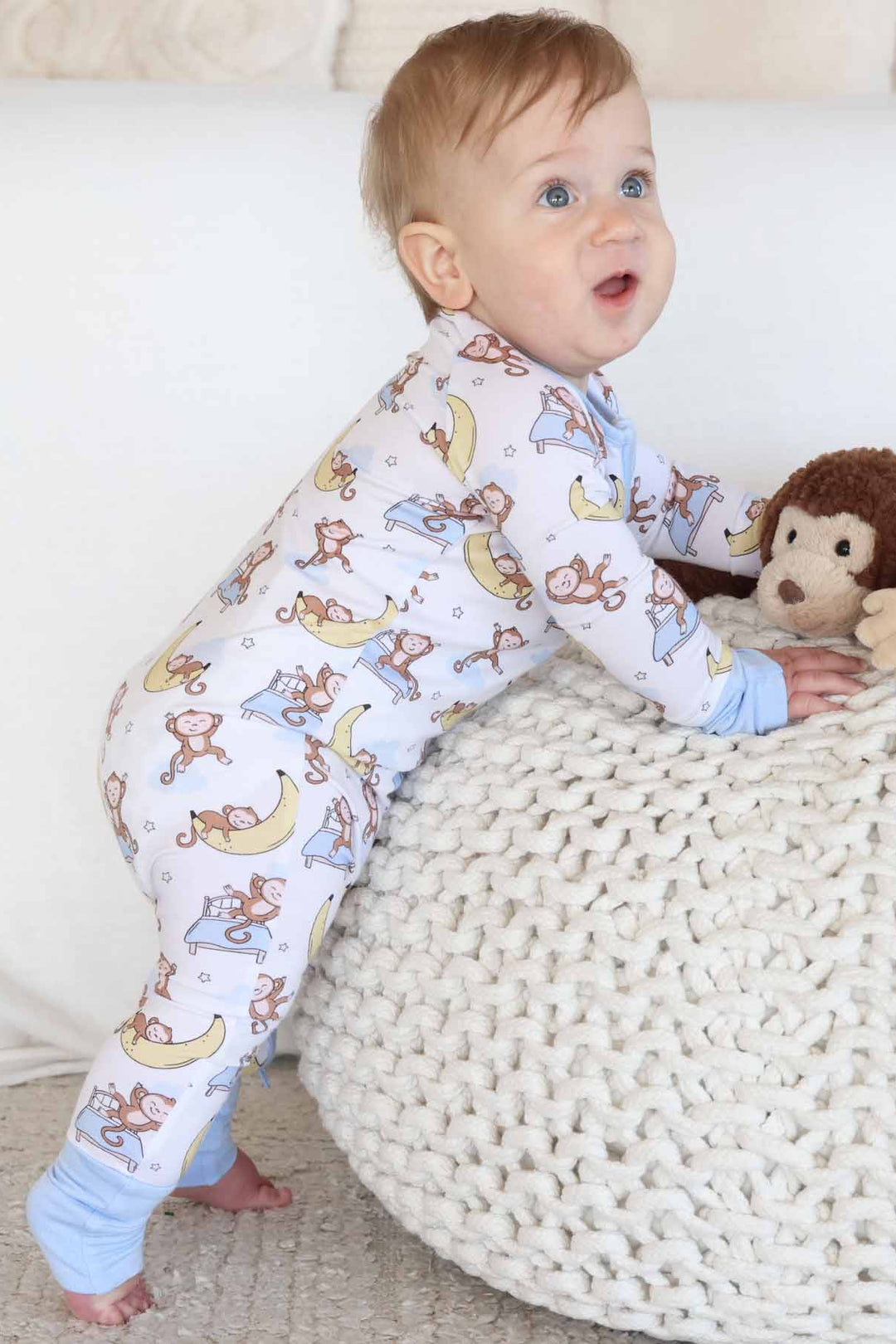 baby wearing monkey and banana convertible zip romper leaning on ottoman with a monkey stuffed animal