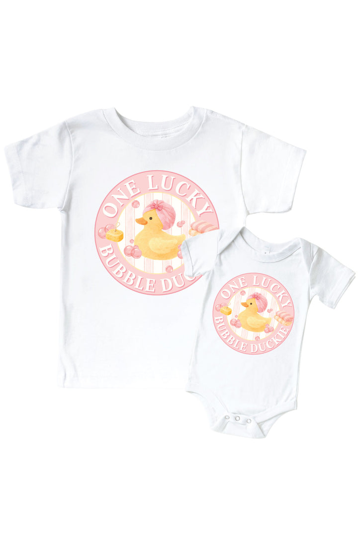 White kids tee and baby bodysuit with pink circle One Lucky Bubble Duckie graphic featuring yellow rubber duck with pink bow and bubbles