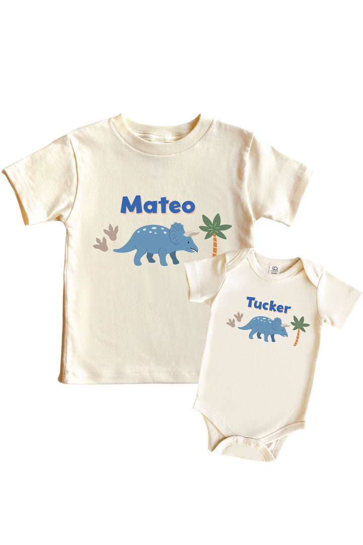 Natural cream kids tee and baby bodysuit set with blue triceratops dinosaur graphic, palm tree accents, and personalized name in blue lettering