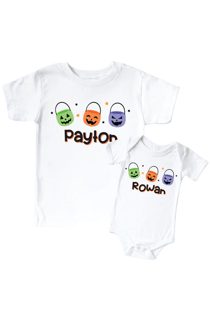 White kids' t-shirt and matching bodysuit personalized with names in orange script below three colorful jack-o-lantern candy bucket icons