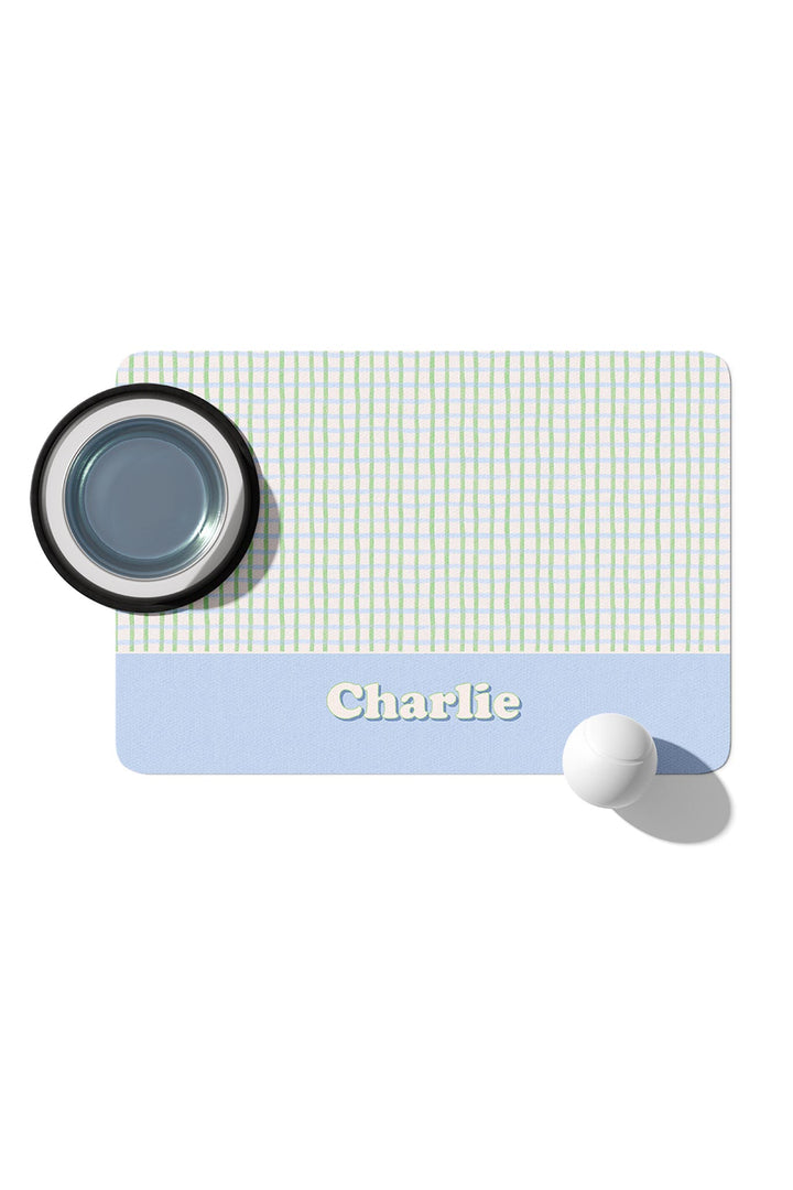 Personalized pet placemat in green and blue picnic plaid with a blue border and the name "Charlie" at the bottom, shown with a pet food bowl on top