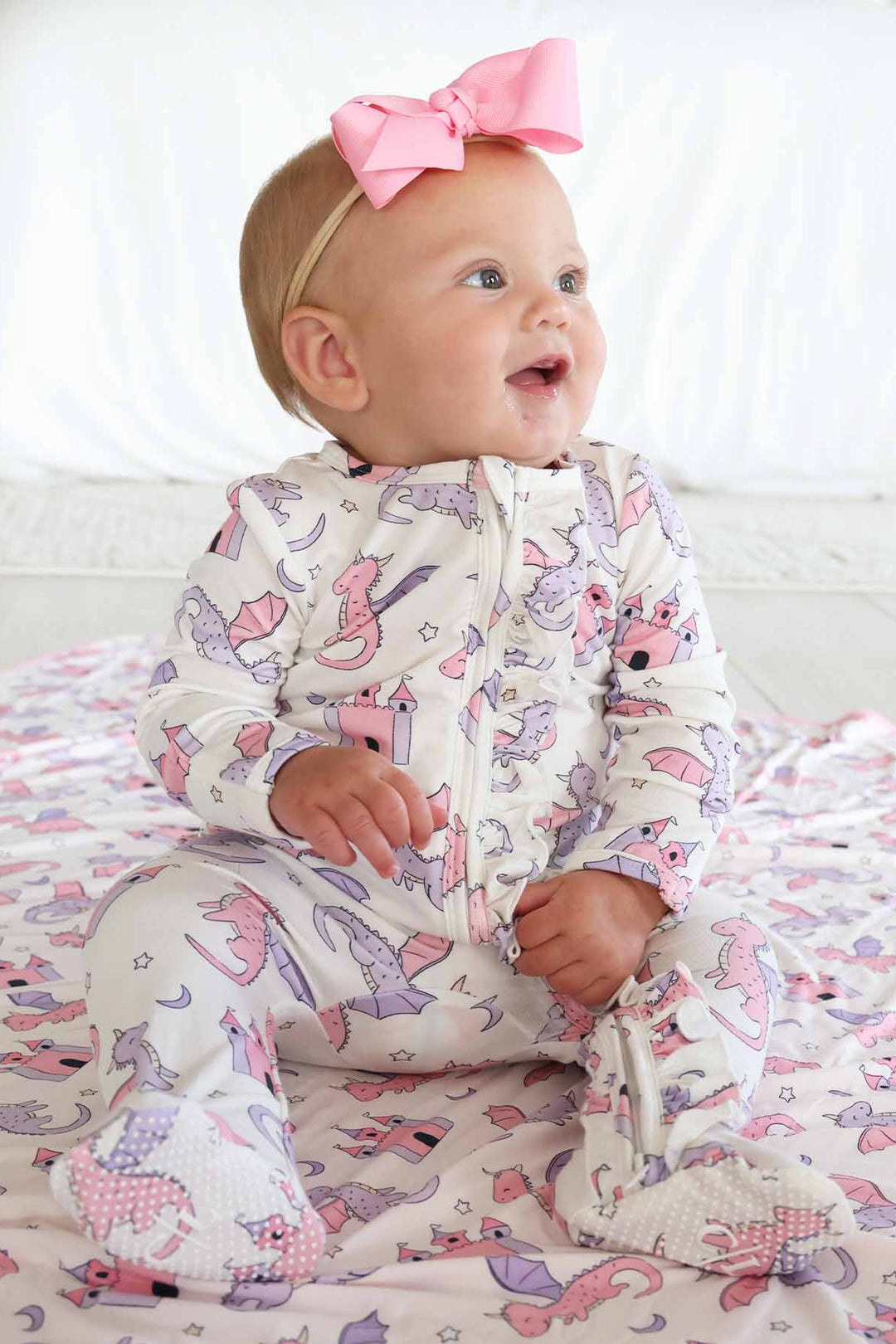 baby girl sitting wearing a pink and purple dragon and castle ruffle zipper footie with pink ribbon bow headband 