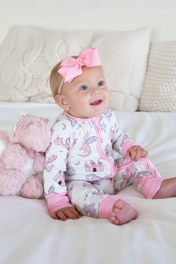 baby girl sitting on bed wearing pink dragon adventures convertible zip romper with matching ribbon bow headband with a pink dragon stuffed animal