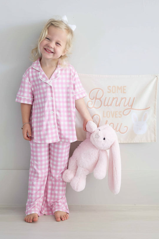 standing girl wearing a short sleeve collared flare lounge set in pink gingham holding a pink stuffed bunny with a some bunny loves you sign behind her 