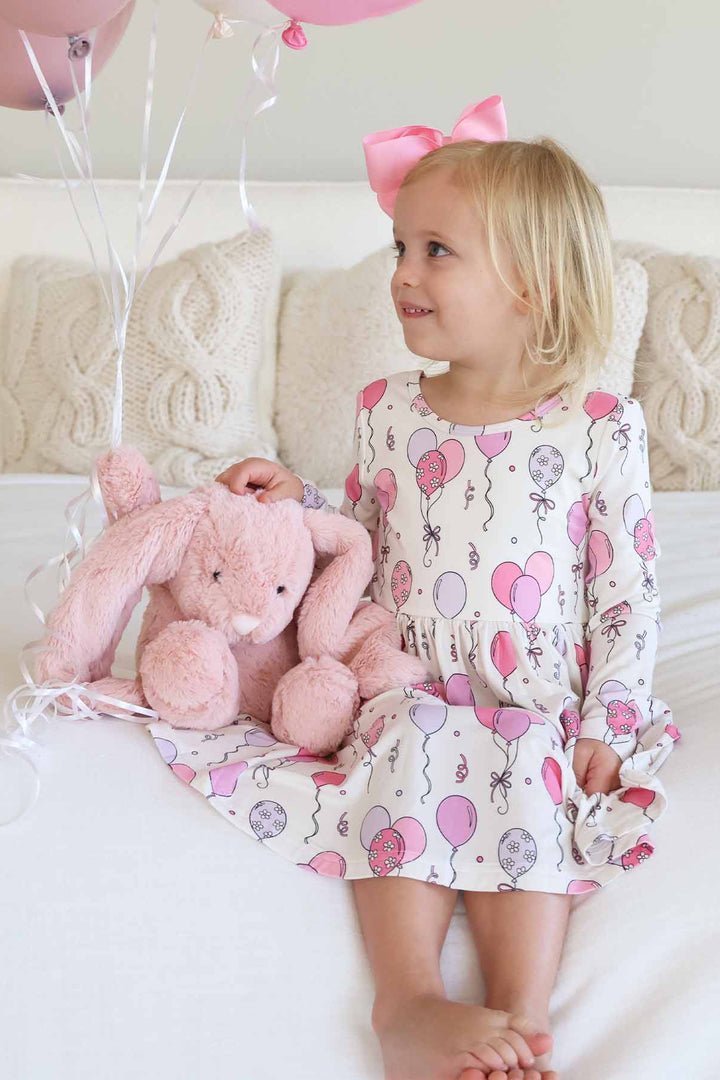 girl sitting on bed wearing pink balloon long sleeve dress next to a pink bunny stuffed animal