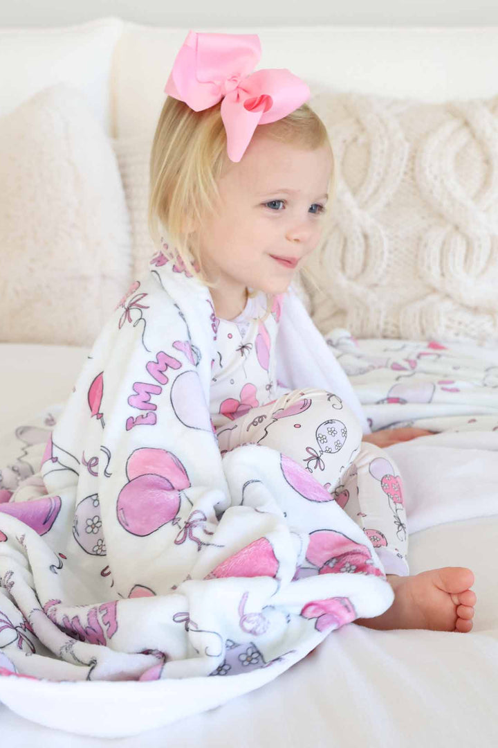 girl sitting on bed with personalized pink balloon and streamer blanket draped over her shoulders 