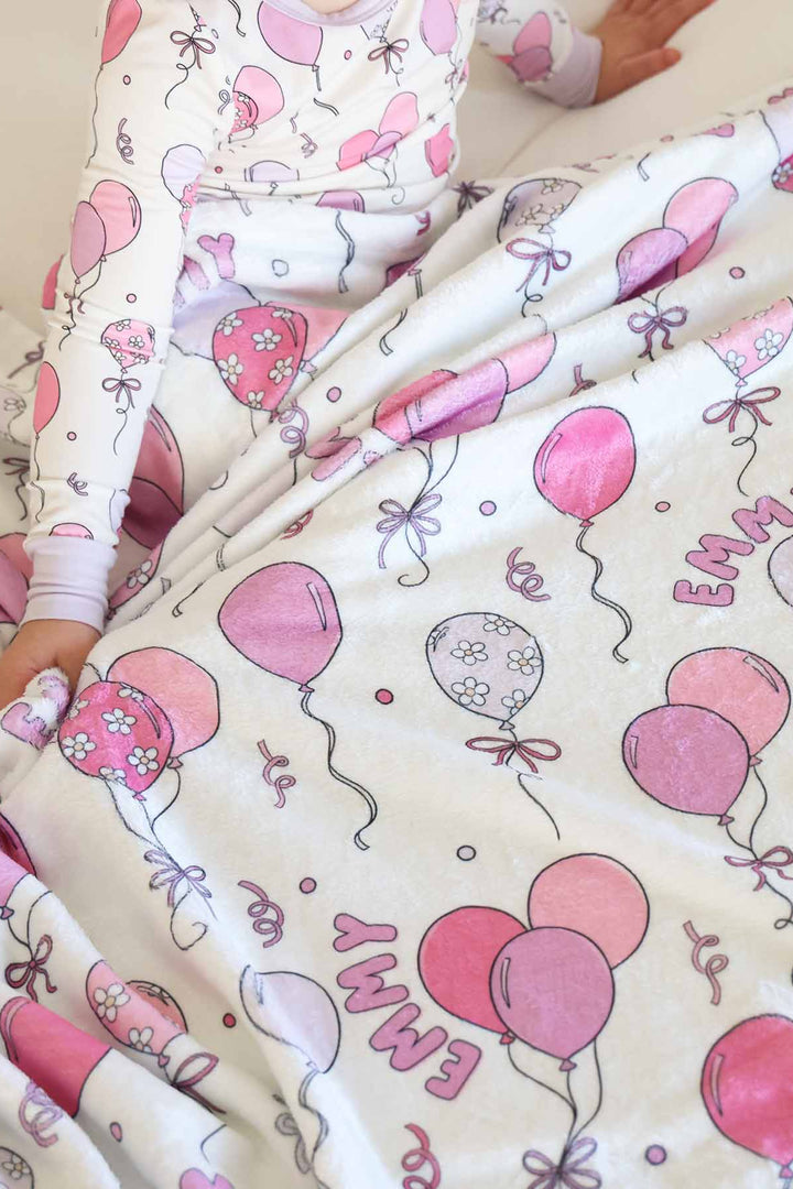 girl sitting on bed wearing a pink balloon long sleeve pajama set covered with a matching personalized blanket 