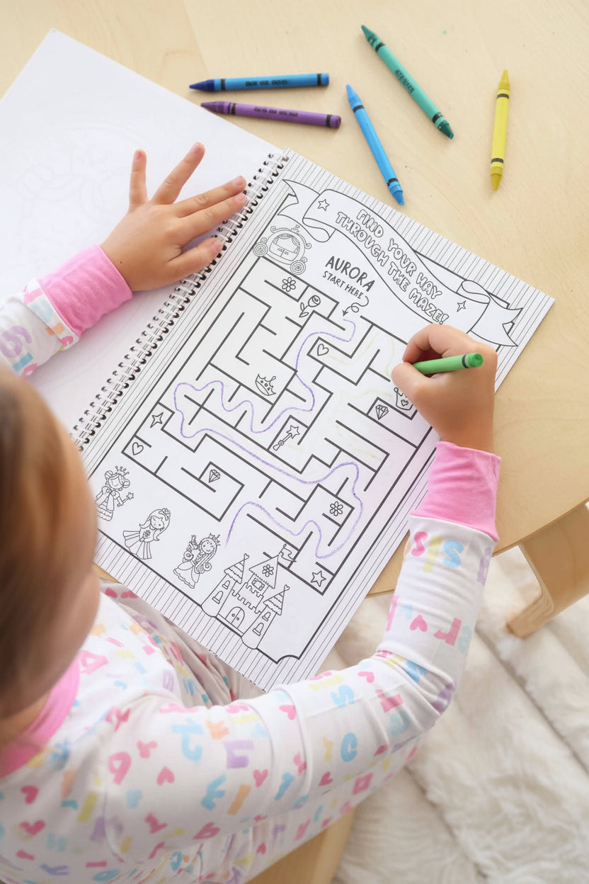 Child in a Big Sister pajama set working through a personalized 'Aurora' maze puzzle page with a green crayon in a Caden Lane coloring book