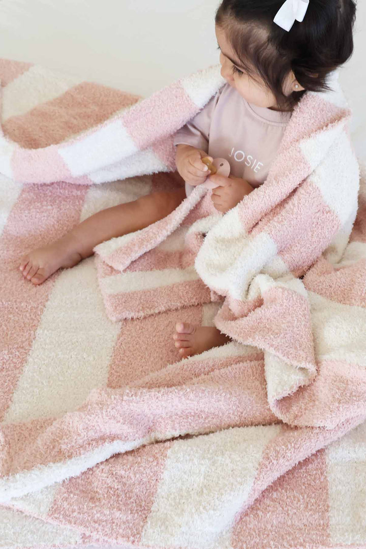 girl sitting on a bed with a pink and white stripe microfiber blanket draped over her holding a matching lovey 