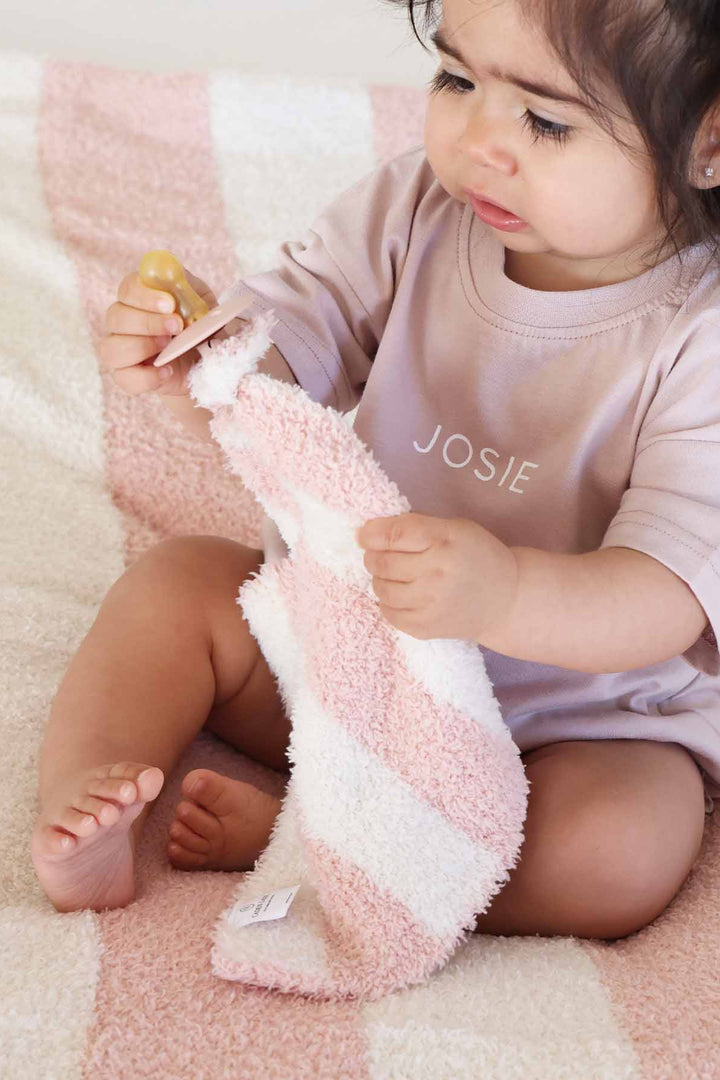 girl sitting on a bed wearing a personalized tshirt romper for babies and toddlers holding a pacifier attached to a pink and white stripe plush lovey and sitting on a matching blanket 