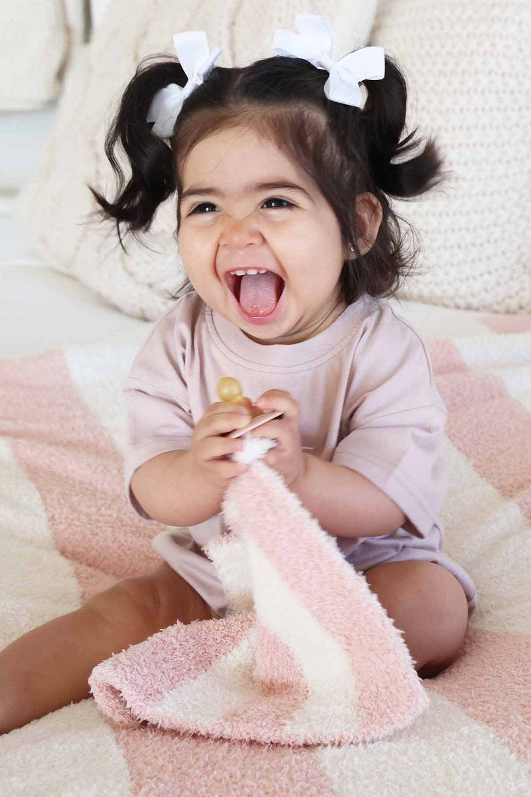 girl sitting on a bed holding a pink and white stripe plush lovey with a pacifier 