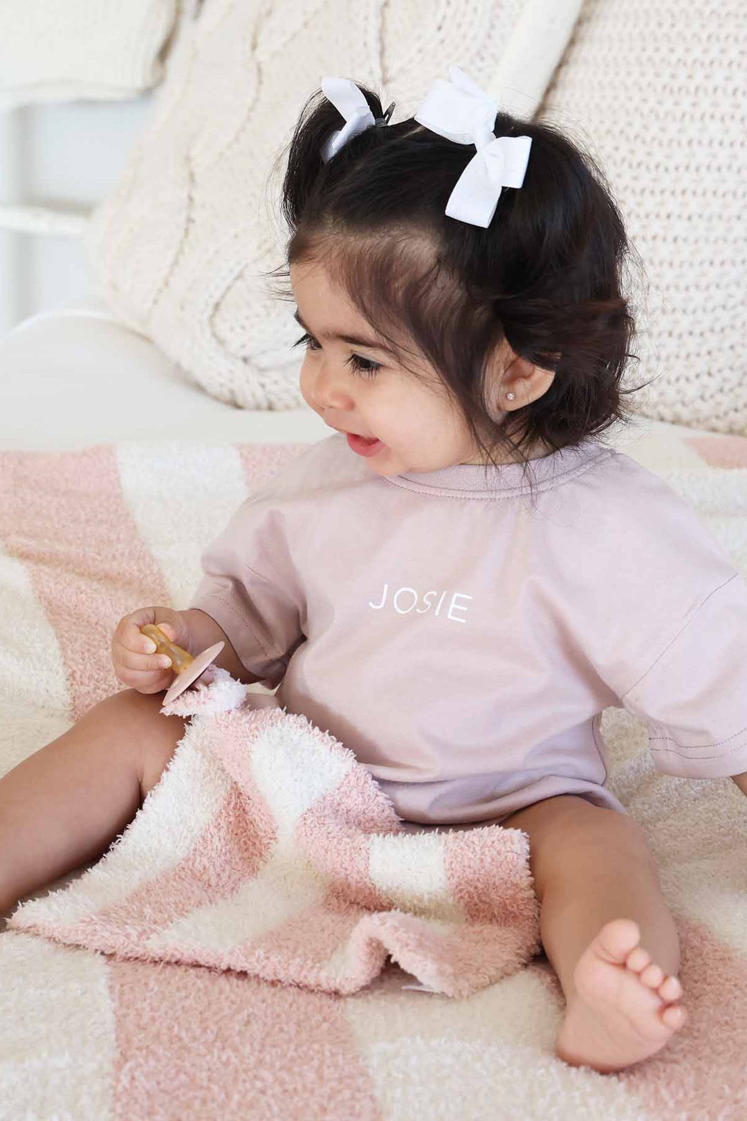 baby girl sitting on a bed wearing a pink and white stripe microfiber blanket holding a matching pink and white stripe lovey