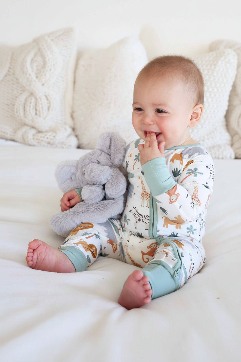 baby sitting on bed in safari pajama romper holding a grey plush elephant lovey