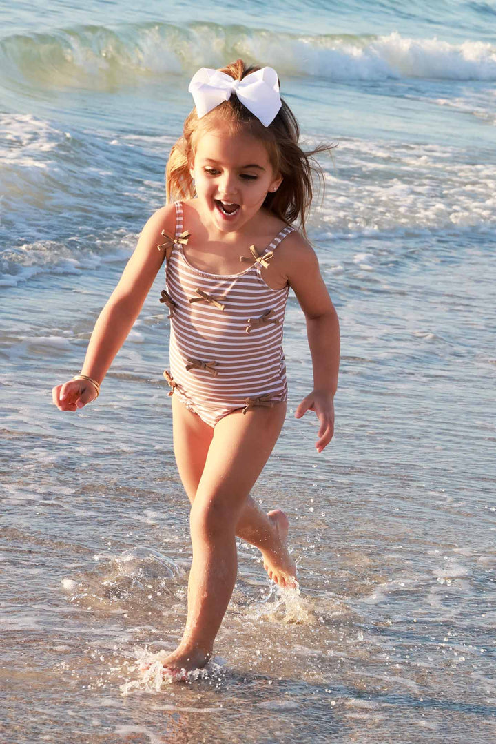 girl running in the water on the beach in a brown and white stripe one piece swimsuit with brown bows on the front 