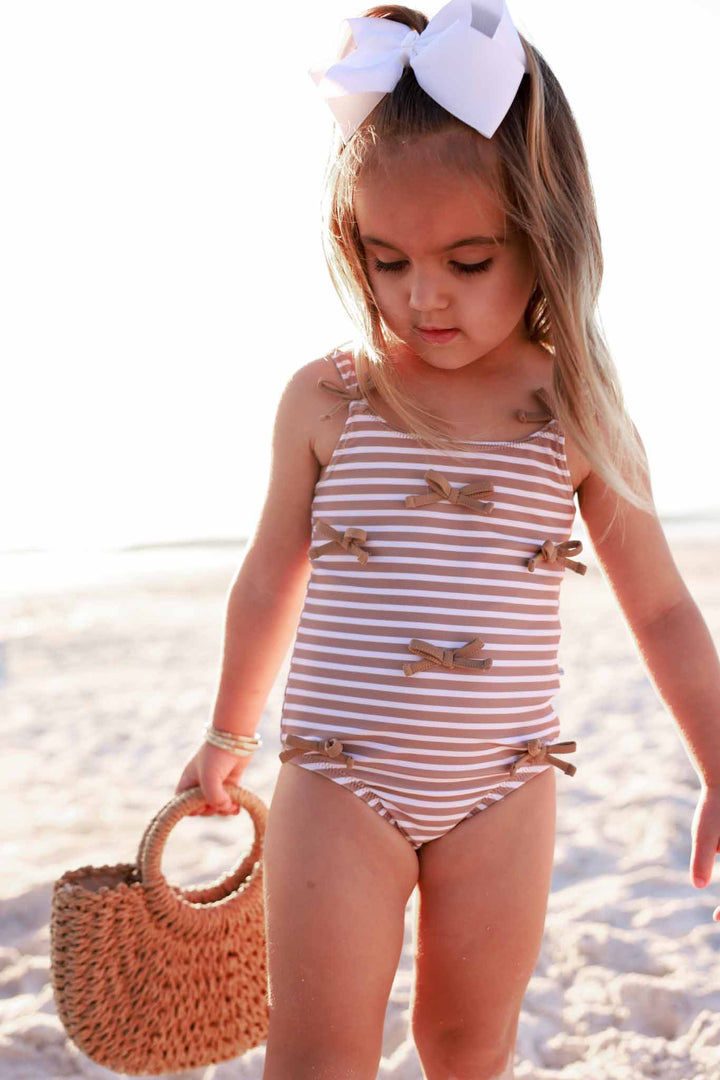 girl standing on beach in brown and white stripe one piece swimsuit with browns bows holding a rattan basket