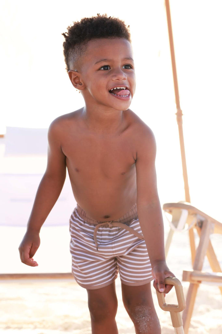 boy playing with a shovel at the beach wearing brown and white stripe swim trunks 