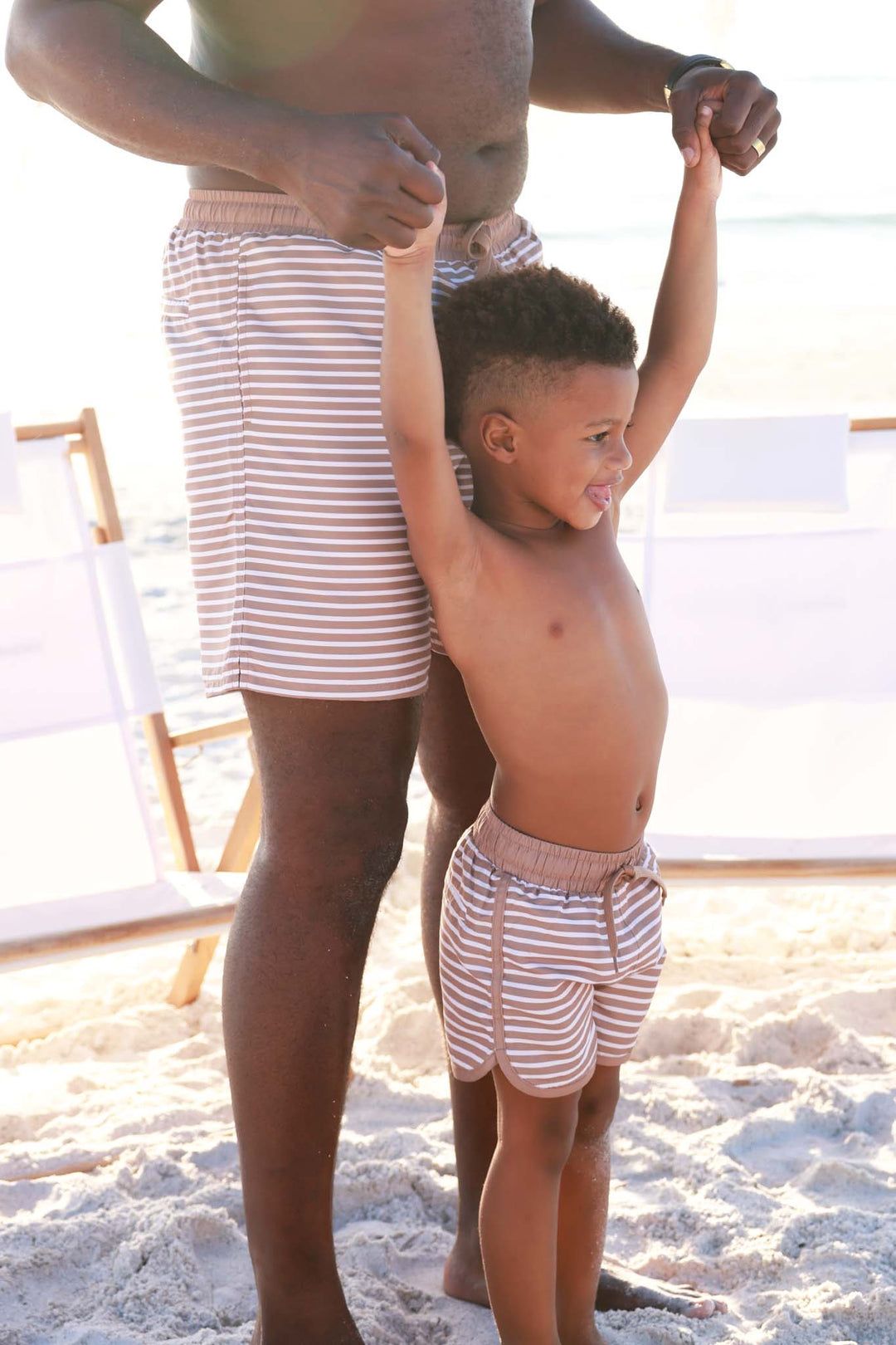 boy standing with dad on the beach in matching brown and white stripe swim trunks 