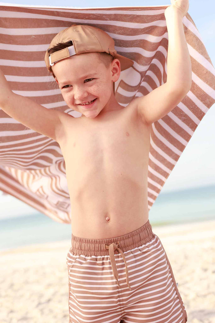 boy at the beach in brown waterproof surf hat with matching striped swim trunks and a personalized towel over his head