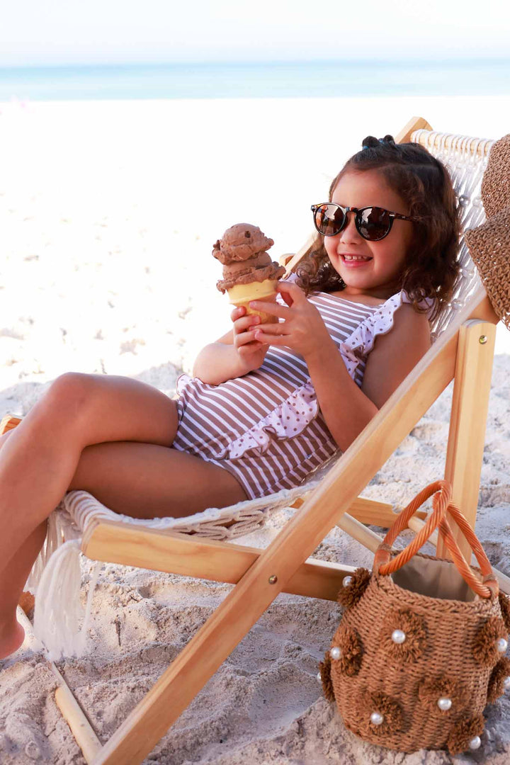 girl laying on beach chair in neutral stripe and polka dot ruffle swimsuit eating a chocolate ice cream cone 