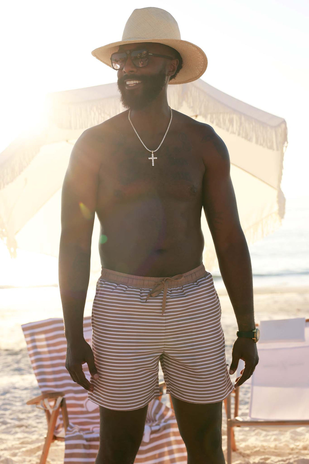 man standing on the beach in neutral stripe swim trunks with sunglasses and a hat 