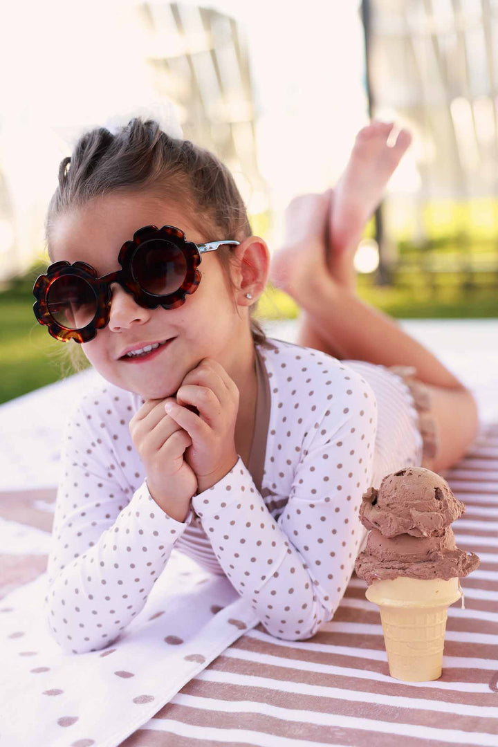 girl laying on towel with polka dot and stripe rash guard romper with tortoise shell sunglasses and a chocolate ice cream cone 