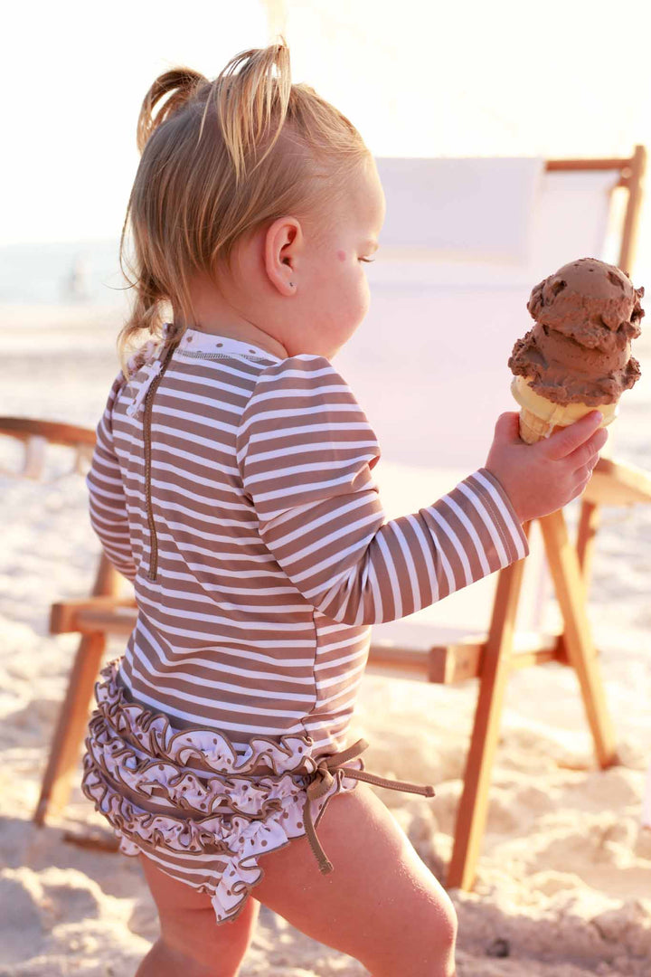baby girl walking away on beach wearing sandy stripes long sleeve rash guard ruffle bottom romper holding a chocolate ice cream cone 