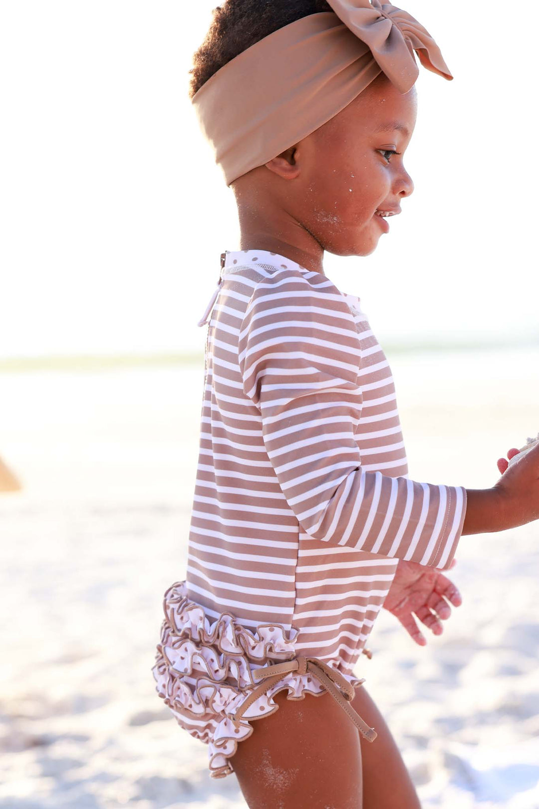 side view of sandy stripes long sleeve ruffle bottom rash guard swimsuit with a matching brown swim bow 