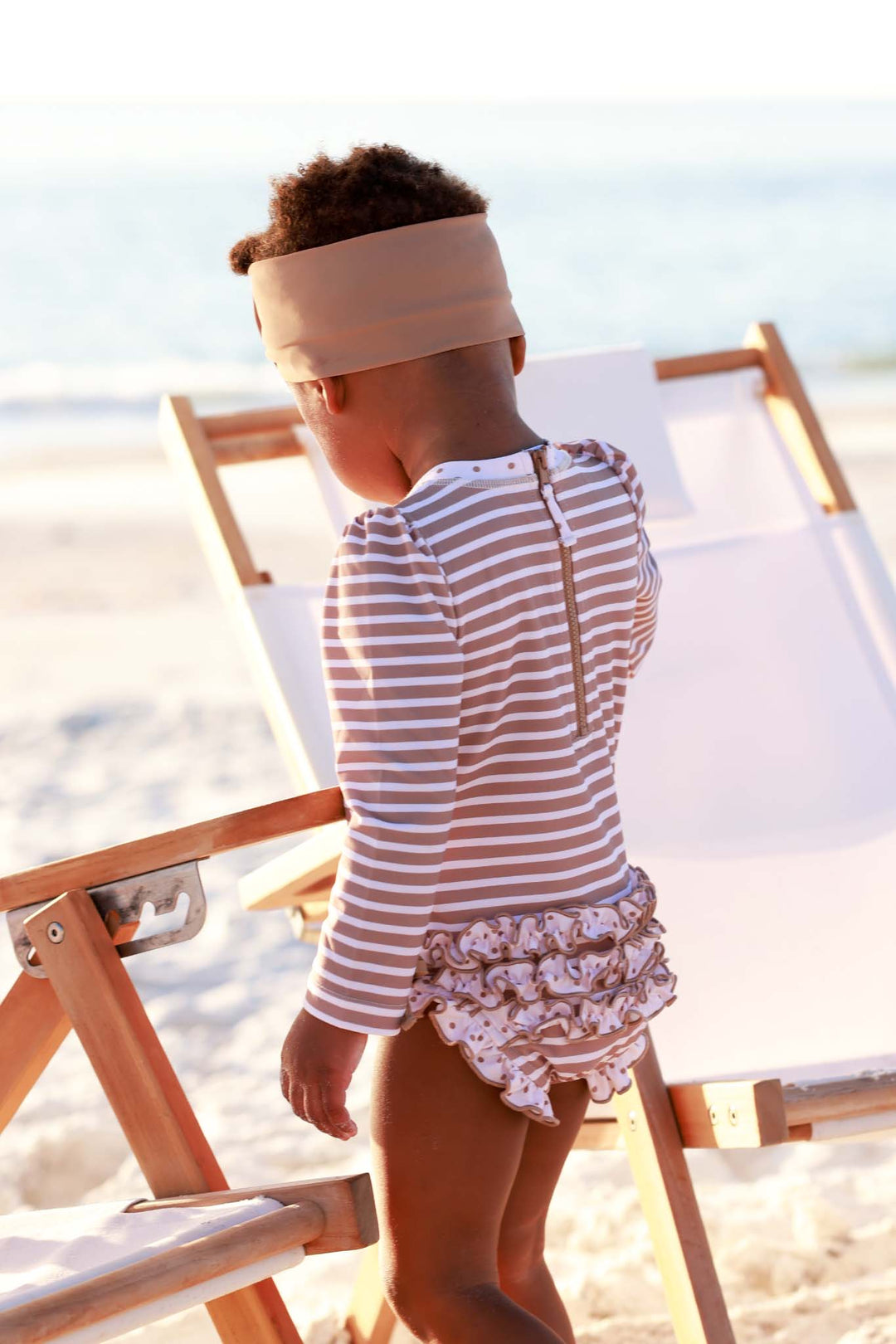 back view of sandy stripes ruffle bottom rash guard swimsuit with brown and white stripes and polka dot accents 