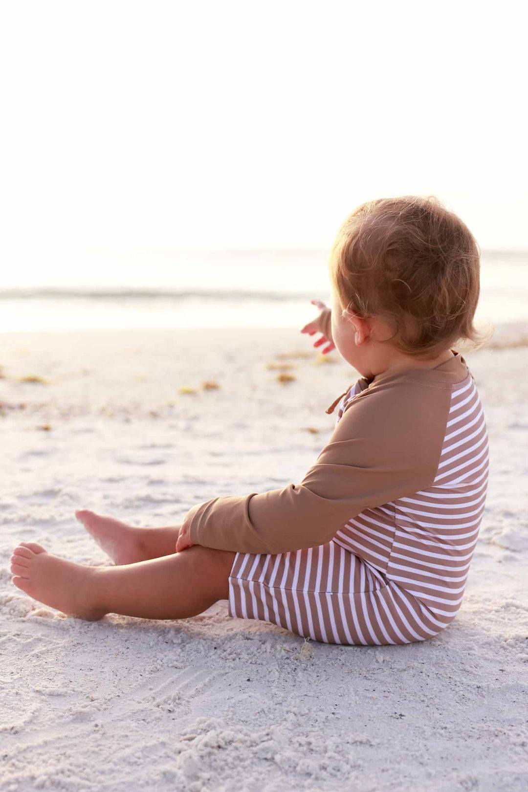 baby boy sitting on beach pointing at ocean in sandy stripes rash guard romper 
