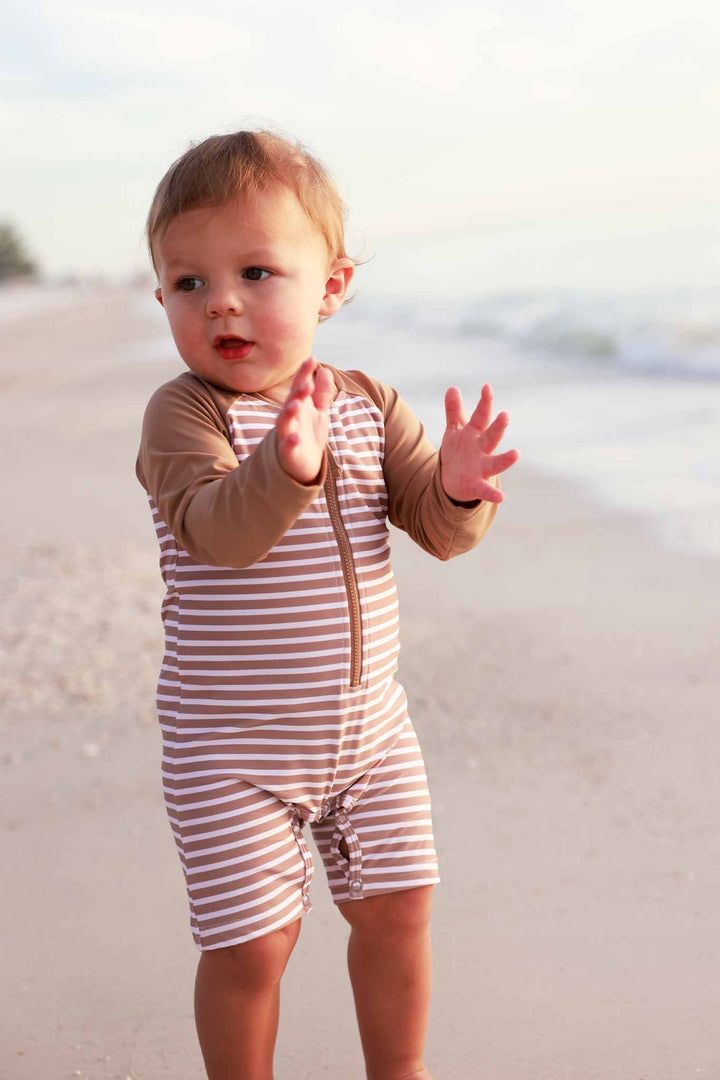 boy standing on beach clapping wearing sandy stripes shorty romper with long sleeves and front zipper 
