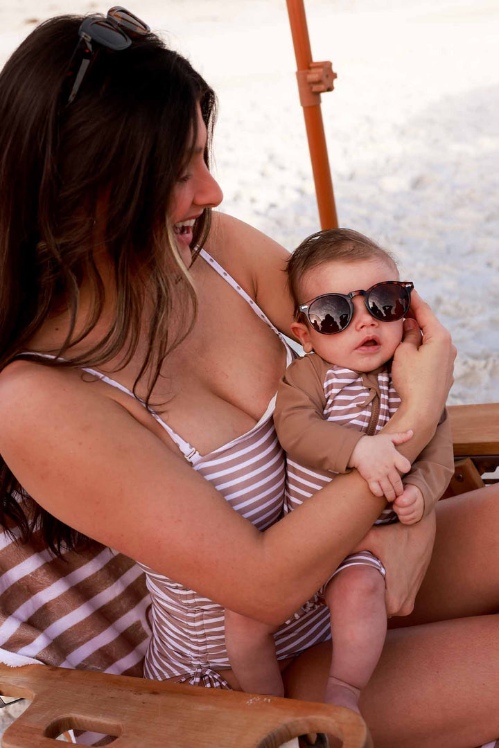 mom and baby wearing matching swimsuits on the beach sitting under an umbrella on a beach chair 
