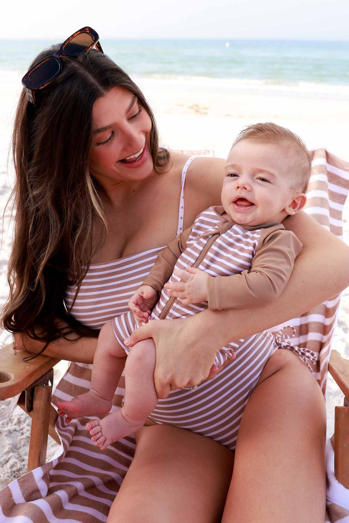 mom holding baby wearing matching sandy stripes swimsuits sitting on a beach chair 