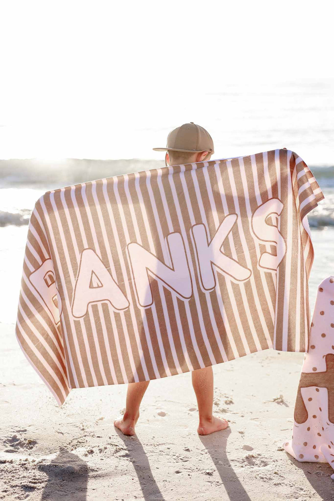 boy standing on the beach with a brown and white stripe personalized towel across his back 